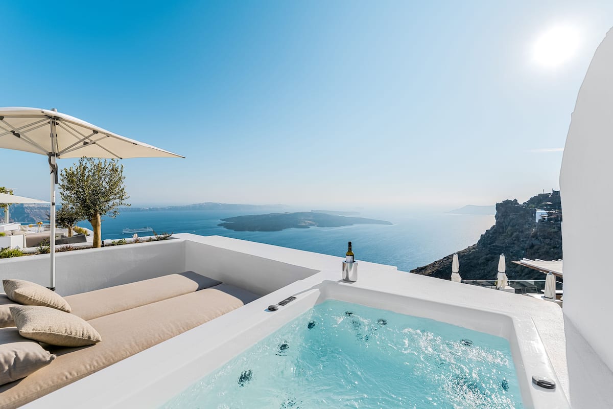 A rooftop infinity pool with a view of the sea, a lounge area with cushions, and a bottle of wine.