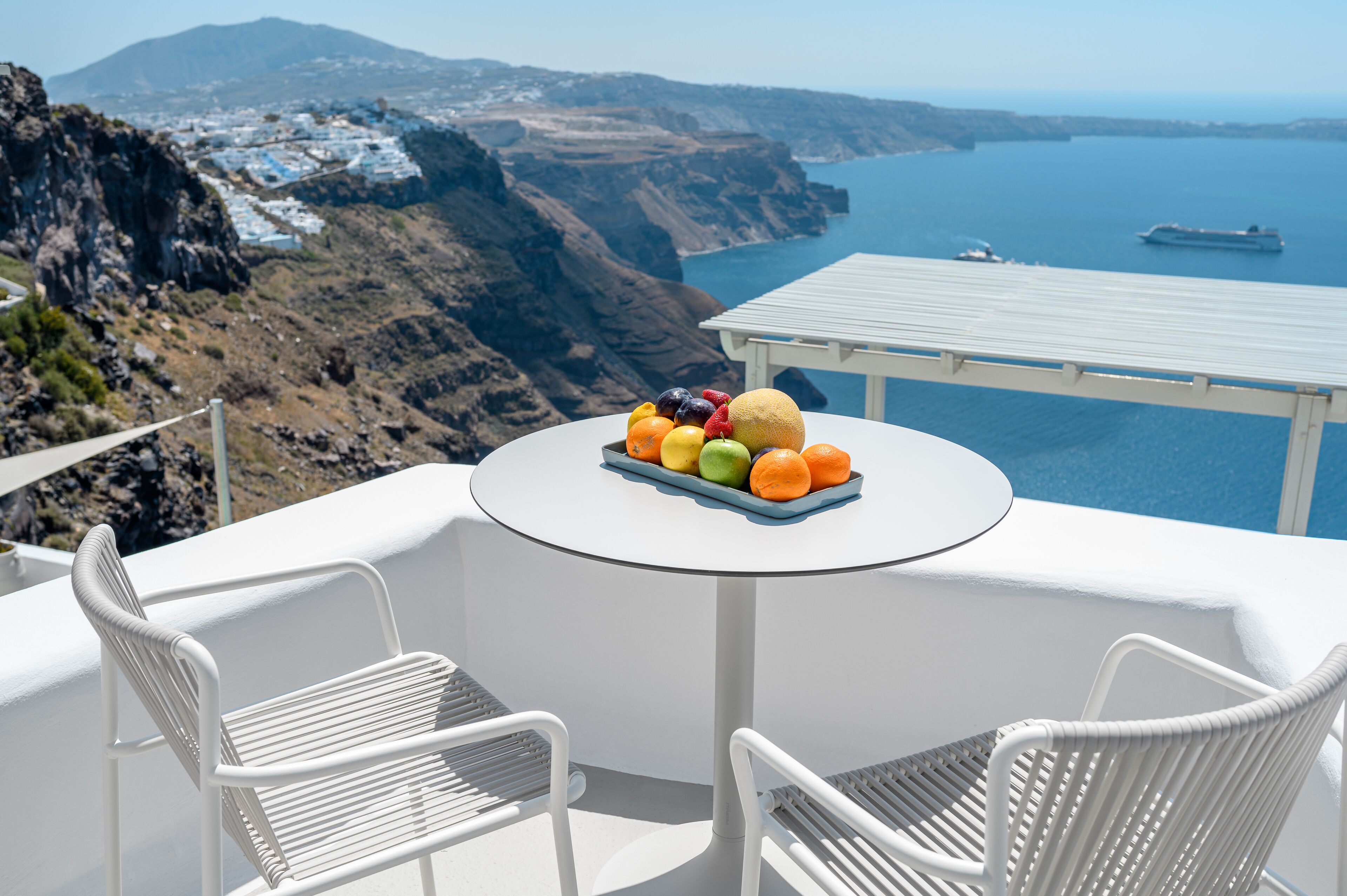 A white outdoor seating area with a table and chairs, overlooking a coastal landscape with cliffs and a body of water.