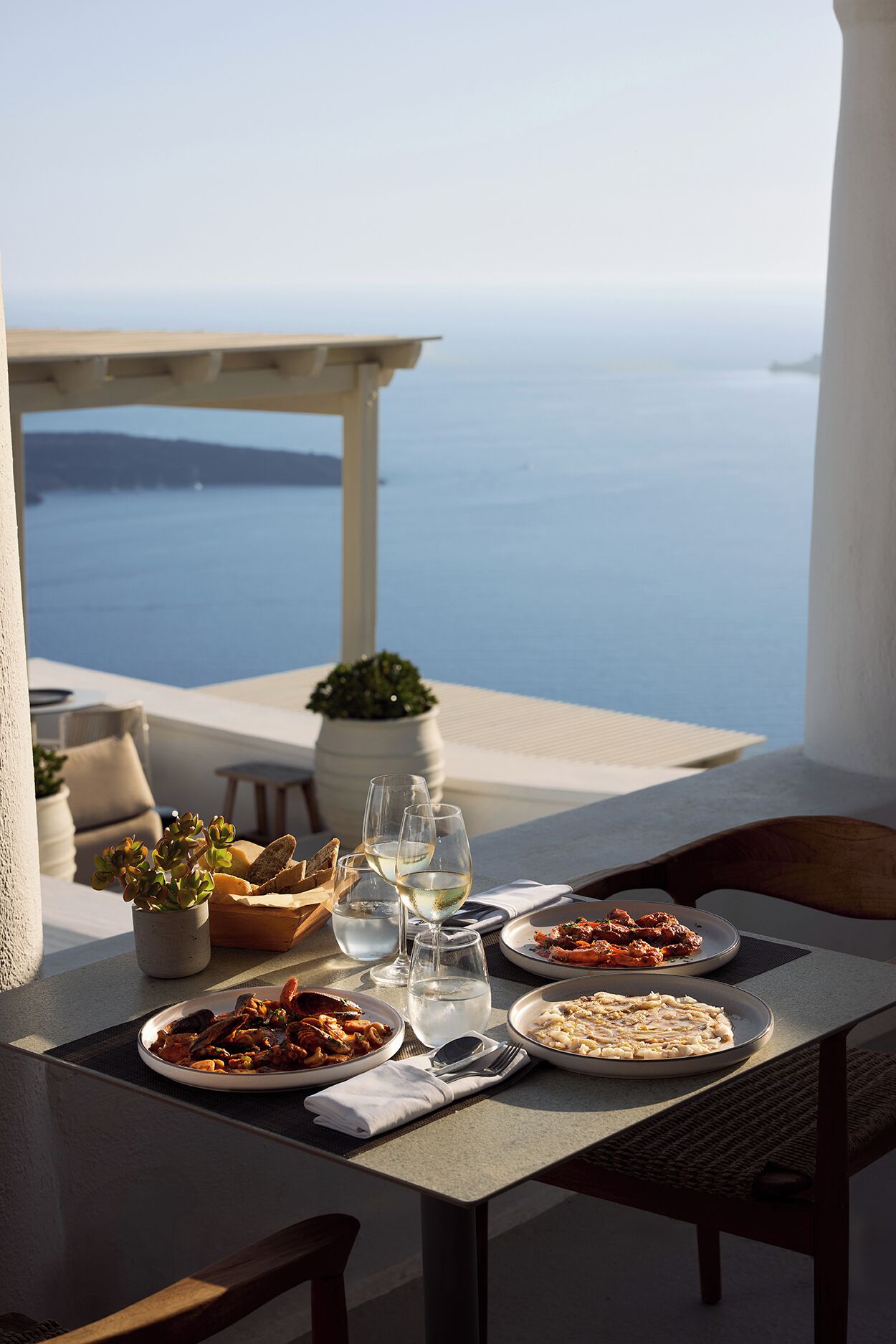 Una mesa lista para una comida, con platos, vasos y una planta en maceta, con vista a un cuerpo de agua.