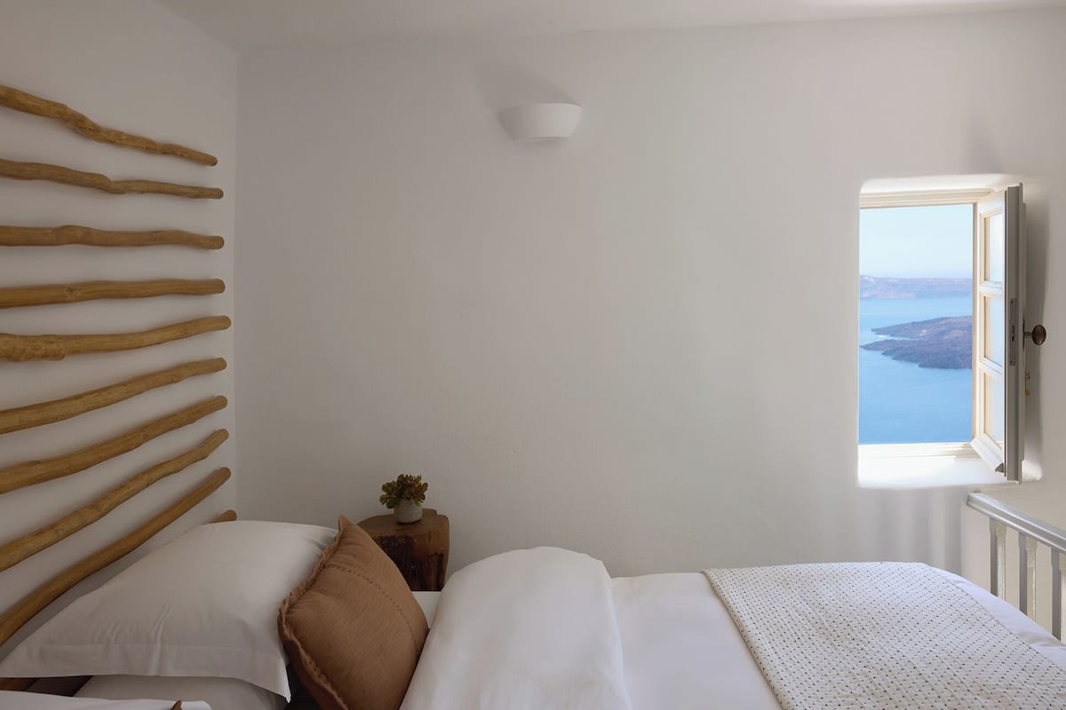 A bedroom with a bed, pillows, a wooden wall art piece, a small bedside table with a plant, and a window with a view of the sea.