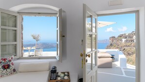 A room with a large window overlooking a scenic coastal view, a cushioned seating area, and a tray with cups and a coffee maker. - Iconic Santorini by Sandglass (Santorini)