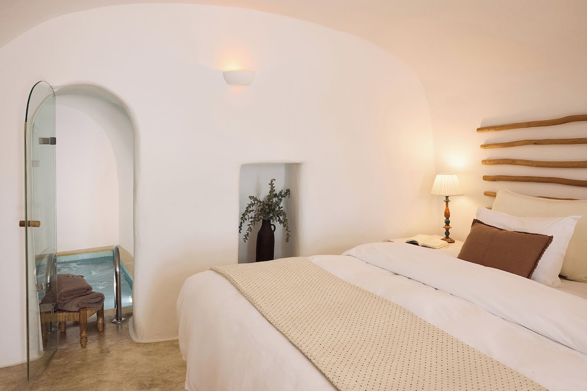 A bedroom with a large bed, a bedside table with a lamp, a glass-enclosed shower area, and a small seating area with a chair and towel.
