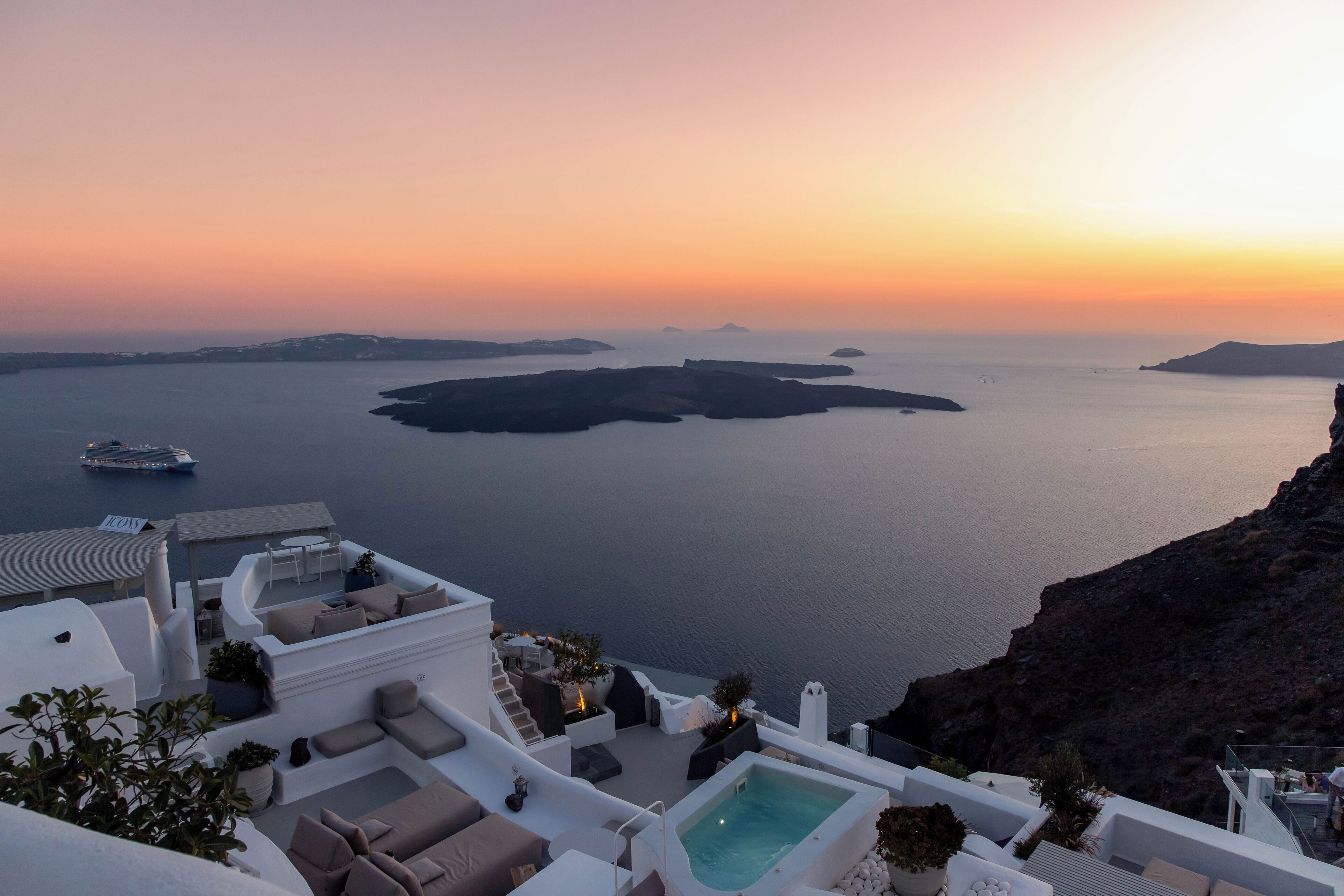 Ein Hotel an der Küste mit Pool, Sitzgelegenheiten im Freien und Blick auf das Meer und die Inseln bei Sonnenuntergang.