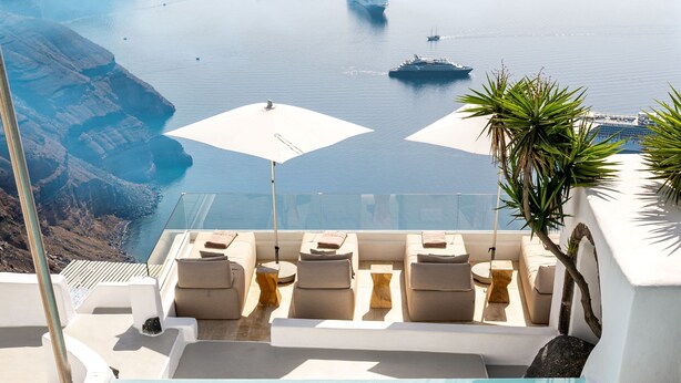 A pool with a person swimming, lounge chairs, and a view of the sea and ships. - Iconic Santorini by Sandglass (Santorini)