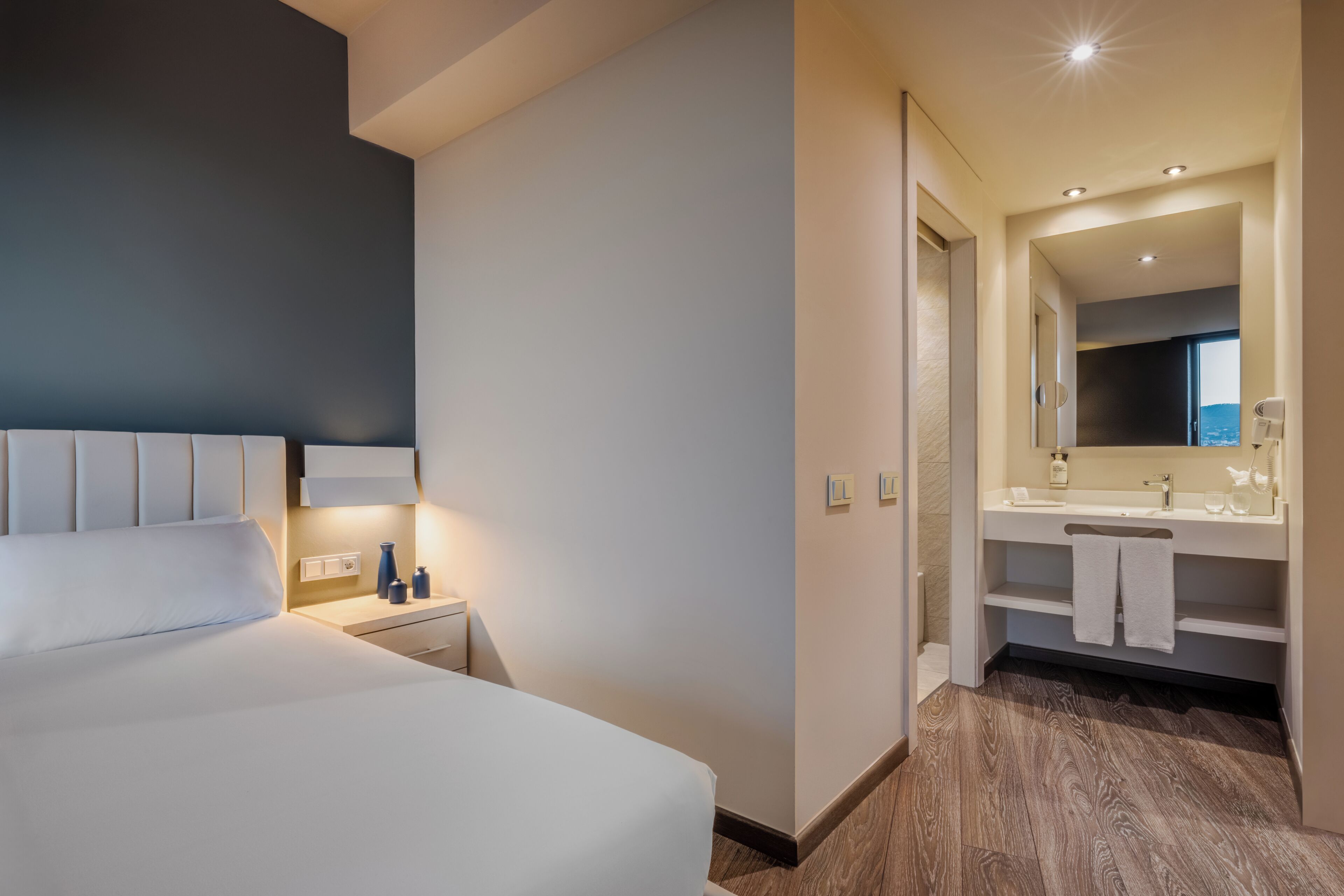 Une chambre d’hôtel comprenant un lit, une table de chevet, une commode et une salle de bain avec un lavabo, un miroir et un porte-serviettes.