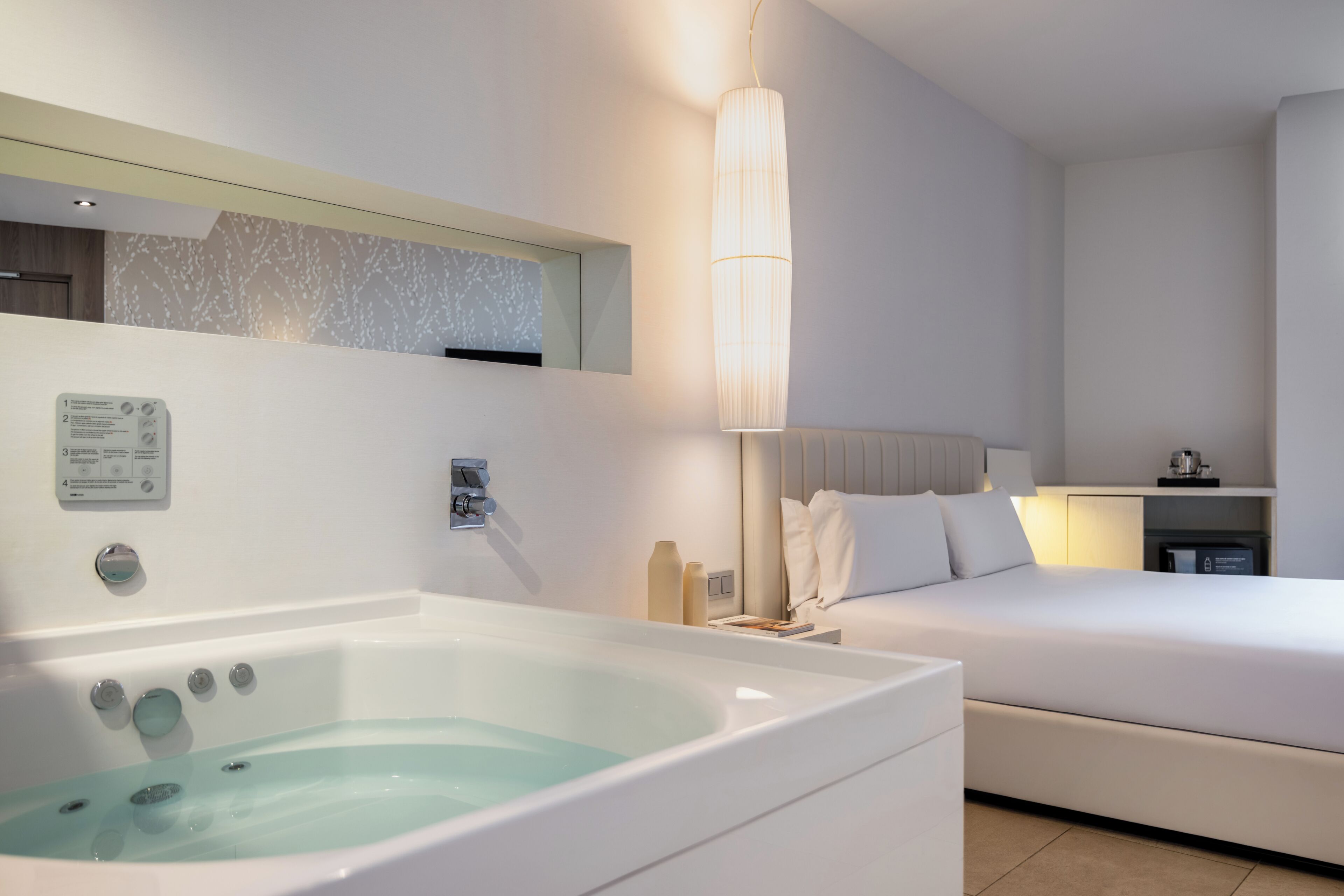 Une chambre d’hôtel moderne équipée d’une grande baignoire, d’un lit, d’une table de chevet et d’un panneau de commande fixé au mur.