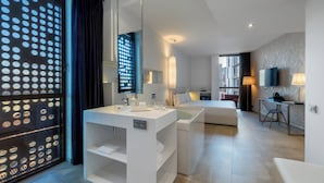 Une chambre d’hôtel moderne avec un grand miroir, un lavabo blanc, une baignoire, un canapé, une télévision et un bureau avec une chaise.