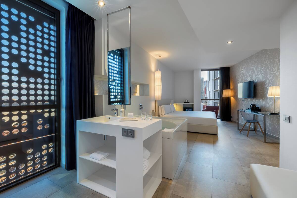 Une chambre d’hôtel moderne avec un grand miroir, un lavabo blanc, une baignoire, un canapé, une télévision et un bureau avec une chaise.