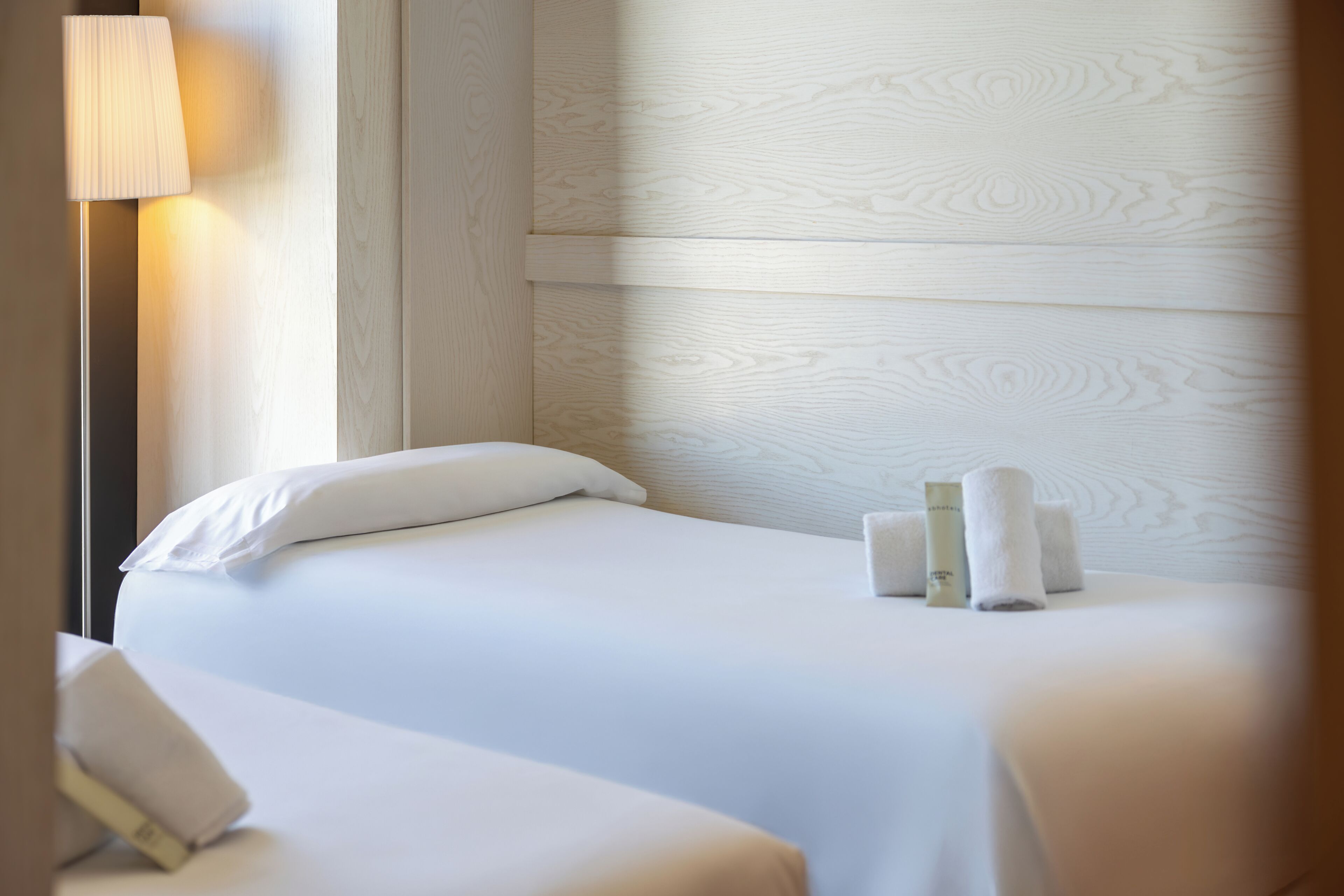 A neatly made bed with pillows, a bedside lamp, and rolled towels on a wooden wall.