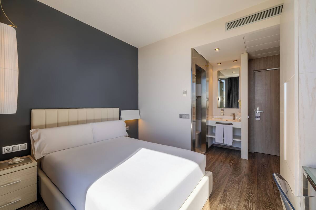 Une chambre d’hôtel moderne dot ée d’un grand lit, de tables de chevet, d’une petite table, d’une salle de bain avec un miroir et d’un espace douche.