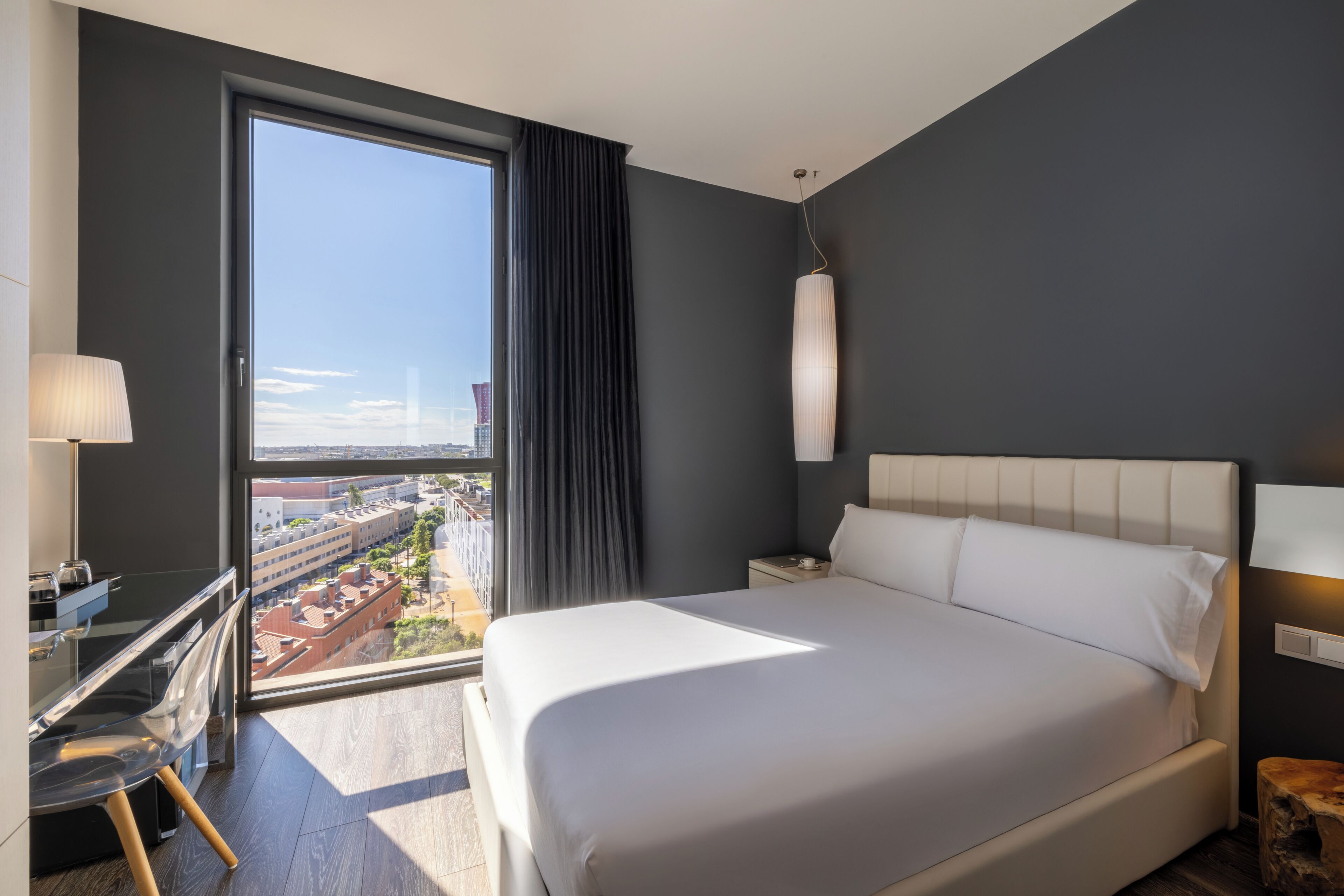 Een moderne hotelkamer met een groot bed, een bureau en uitzicht op de stad door een groot raam.