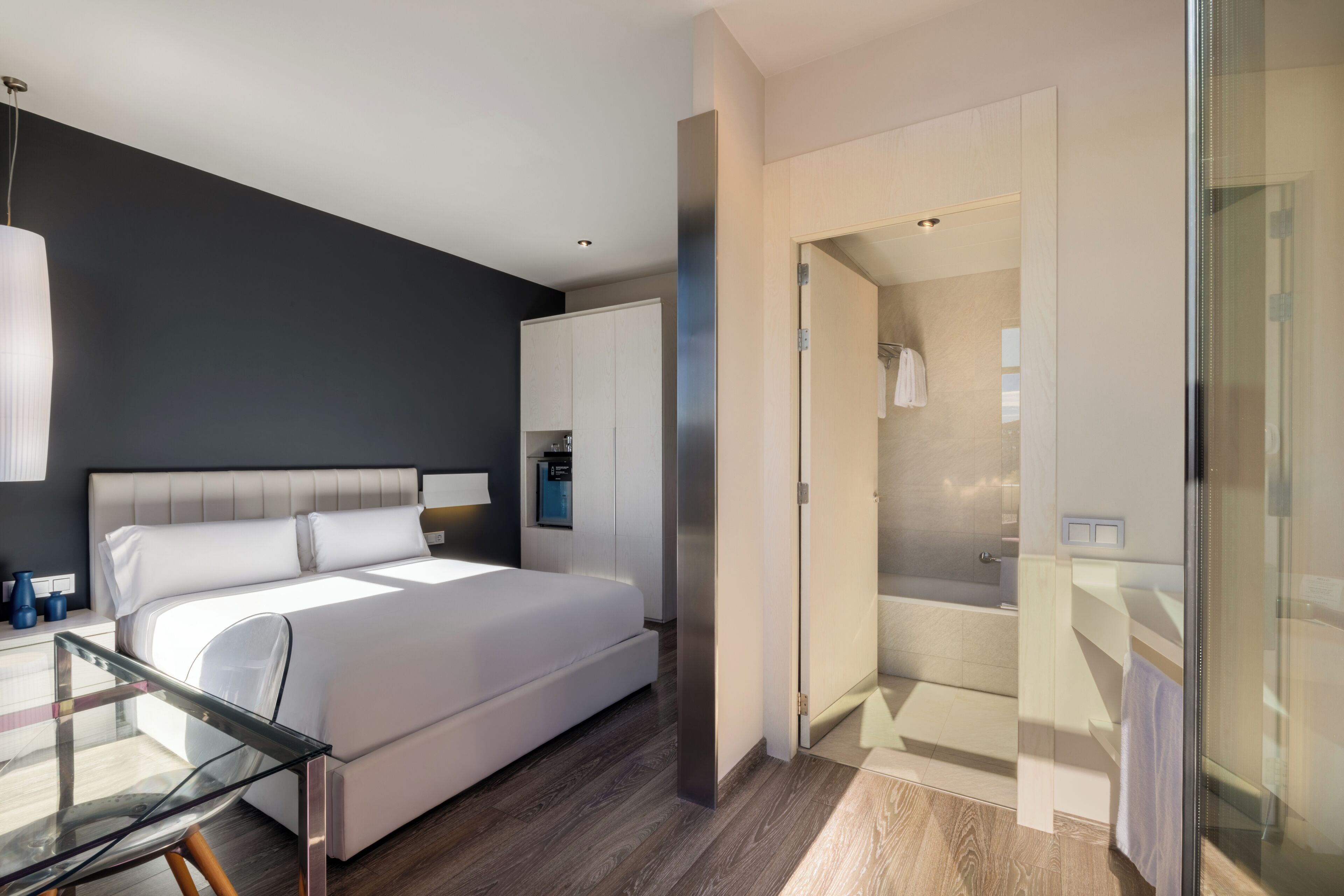 A modern hotel room with a large bed, a glass table, a walk-in closet, and a glass-enclosed shower area.