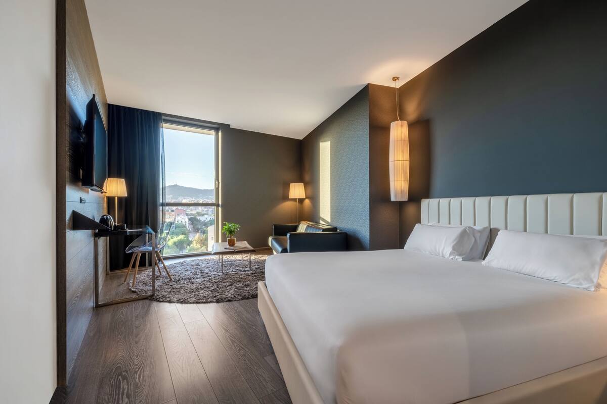 Une chambre d’hôtel moderne dotée d’un grand lit, d’un bureau avec une chaise, d’un téléviseur fixé au mur et d’une fenêtre donnant sur la ville.