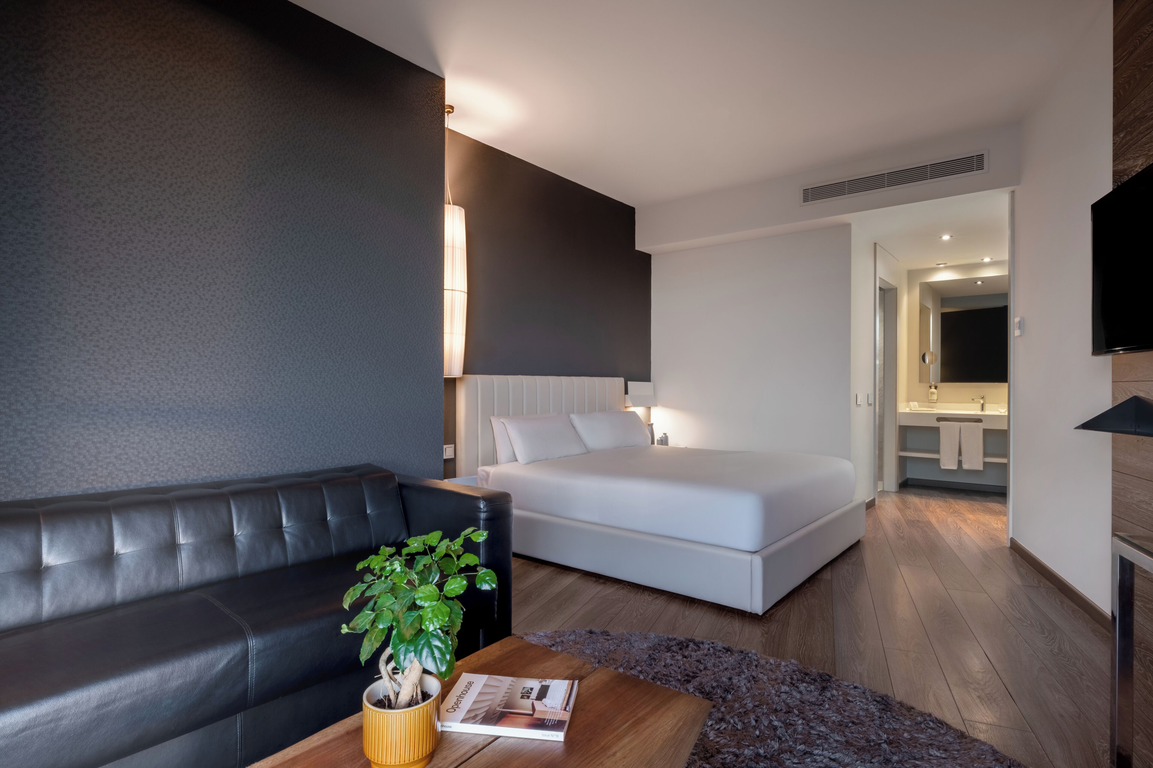 Une chambre d’hôtel moderne avec un canapé en cuir noir, une table basse en bois agrémentée d’une plante, un lit blanc et un téléviseur fixé au mur.