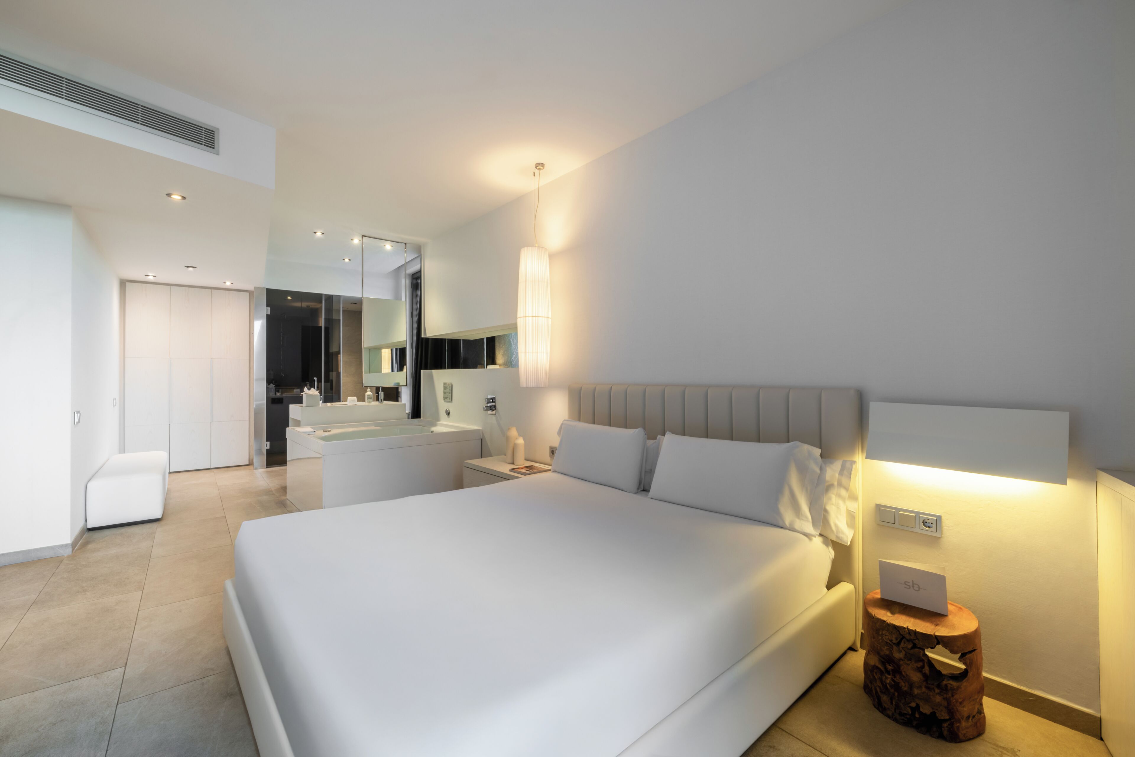 Una habitación de hotel moderna con una cama grande, una mesita de noche, una pequeña mesa auxiliar de madera, un espejo y un baño a la vista al fondo.