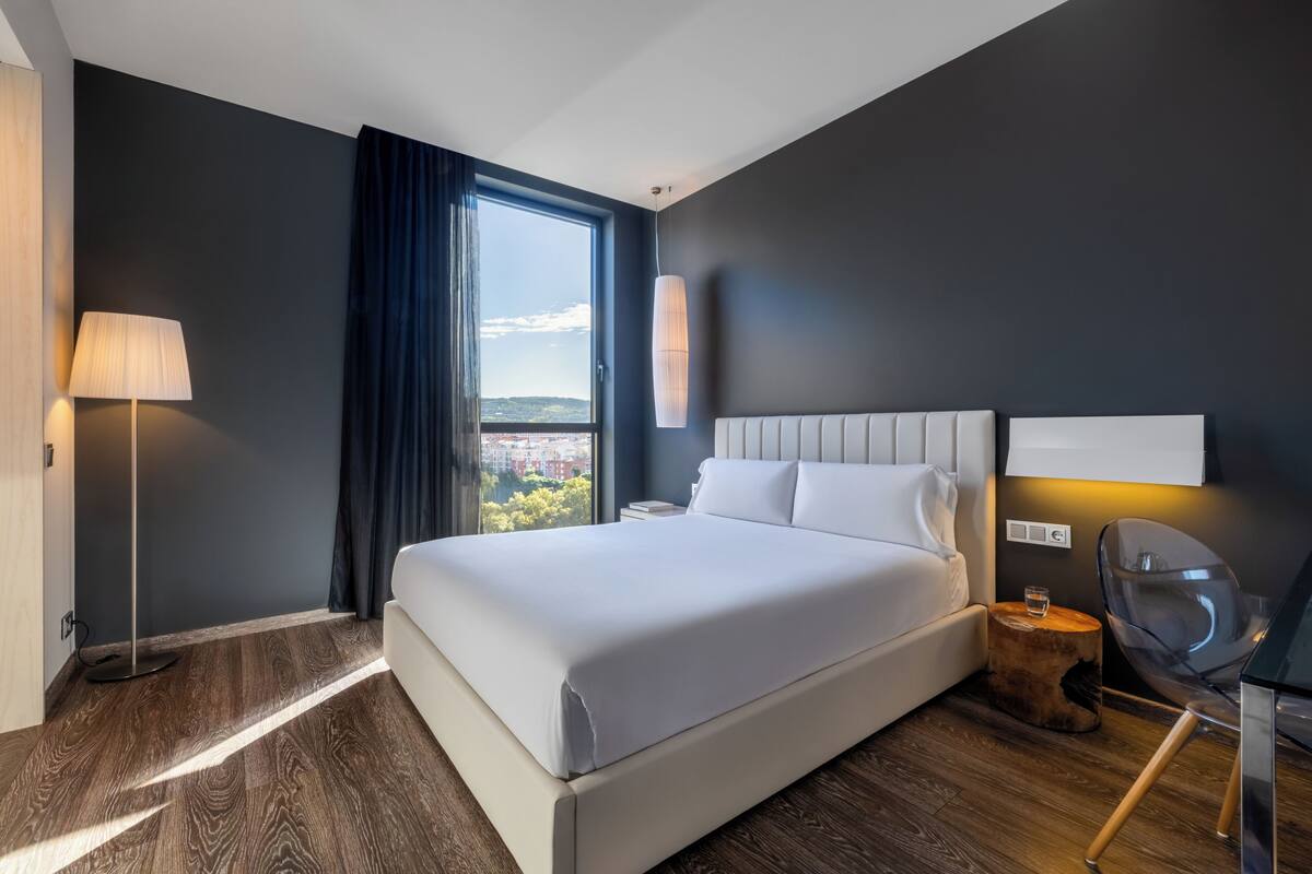 Une chambre d’hôtel moderne avec un grand lit, une table de chevet, une lampe, une chaise et une vue sur la ville.