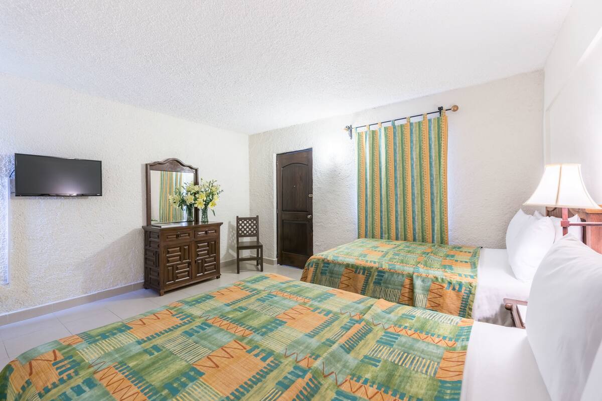 Ein Schlafzimmer mit zwei Betten, einem Schminktisch, einem Fernseher, einem Sessel und einem Fenster mit Vorhängen.