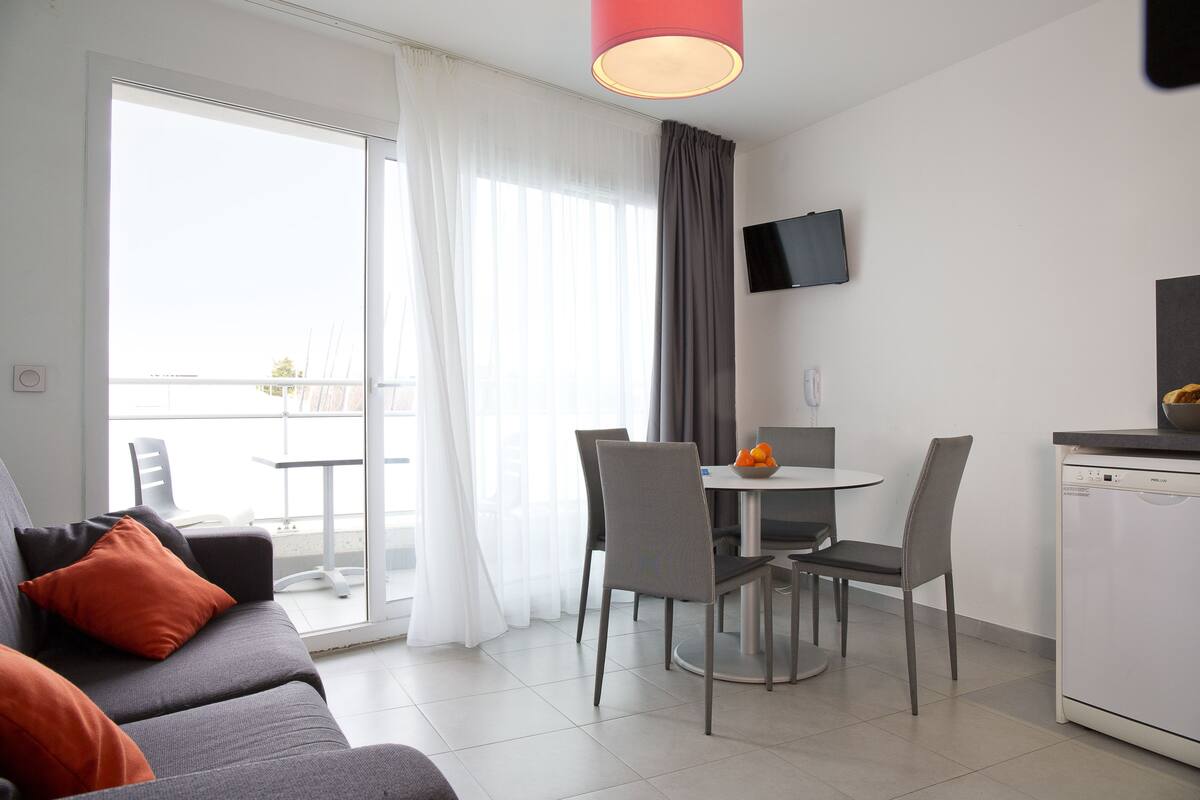 Ein modernes Wohnzimmer mit einem grauen Sofa, orangefarbenen Kissen, einem Esstisch mit Stühlen, einem Flachbildfernseher und einem Geschirrspüler.