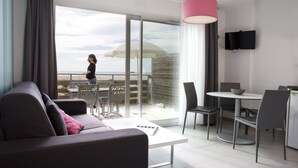 A modern living room with a sofa, a dining table with chairs, a TV mounted on the wall, and a balcony with a view of the beach. - Residence Côté Mer (La Grande-Motte)