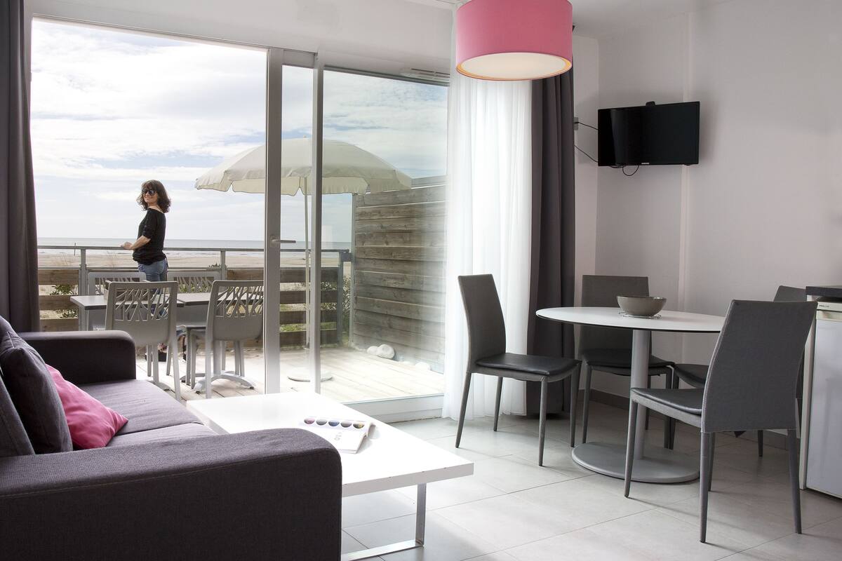 Ein modernes Wohnzimmer mit einer Couch, einem Esstisch mit Stühlen, einem an der Wand befestigten Fernseher und einem Balkon mit Blick auf den Strand.