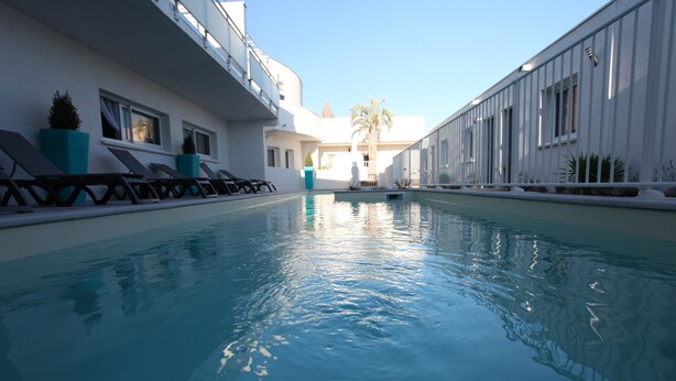 A swimming pool with lounge chairs and potted plants, surrounded by a white building with large windows. - Residence Côté Mer (La Grande-Motte)