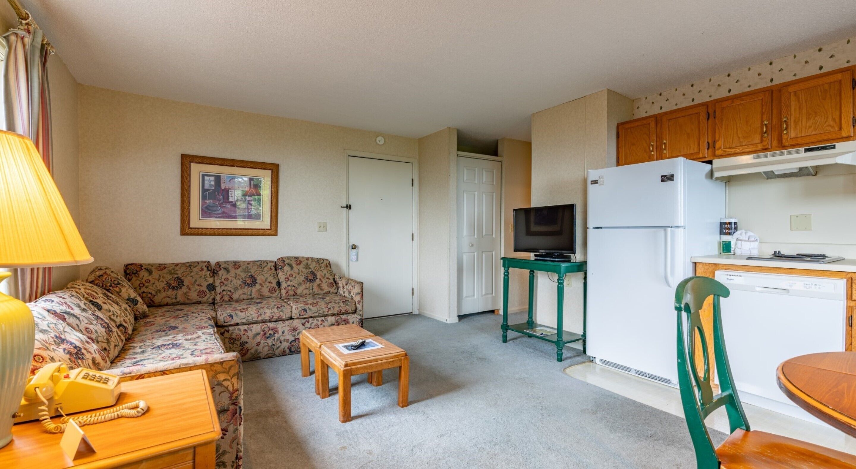 A compact living space with a sofa, a small wooden table, a television on a stand, a refrigerator, and a kitchen area with wooden cabinets.