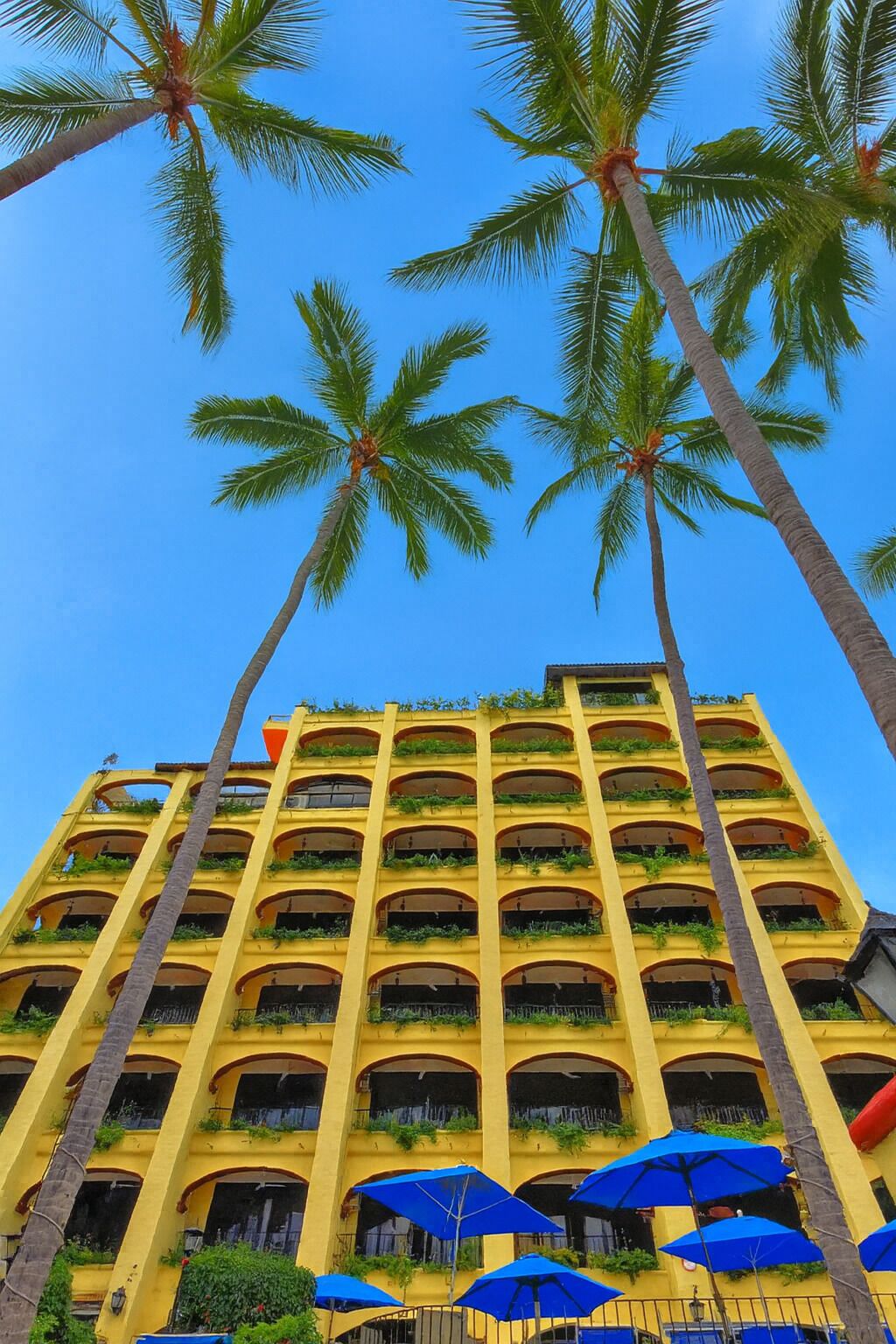 Un hotel de varios pisos con balcones, sombrillas azules y palmeras.