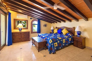 Quarto com cama, duas mesas de cabeceira, cômoda, ventilador de teto e janela com cortinas.