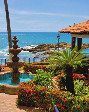 A pool area with a tiered fountain, surrounded by lush greenery and red flowers, overlooking a rocky coastline. - Lindo Mar Resort (Puerto Vallarta)