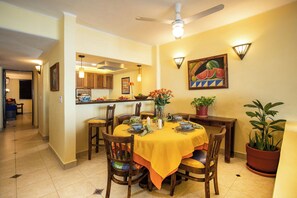 A dining area with a table set for four, surrounded by wooden chairs, a ceiling fan, and a kitchen in the background. - Lindo Mar Resort (Puerto Vallarta)