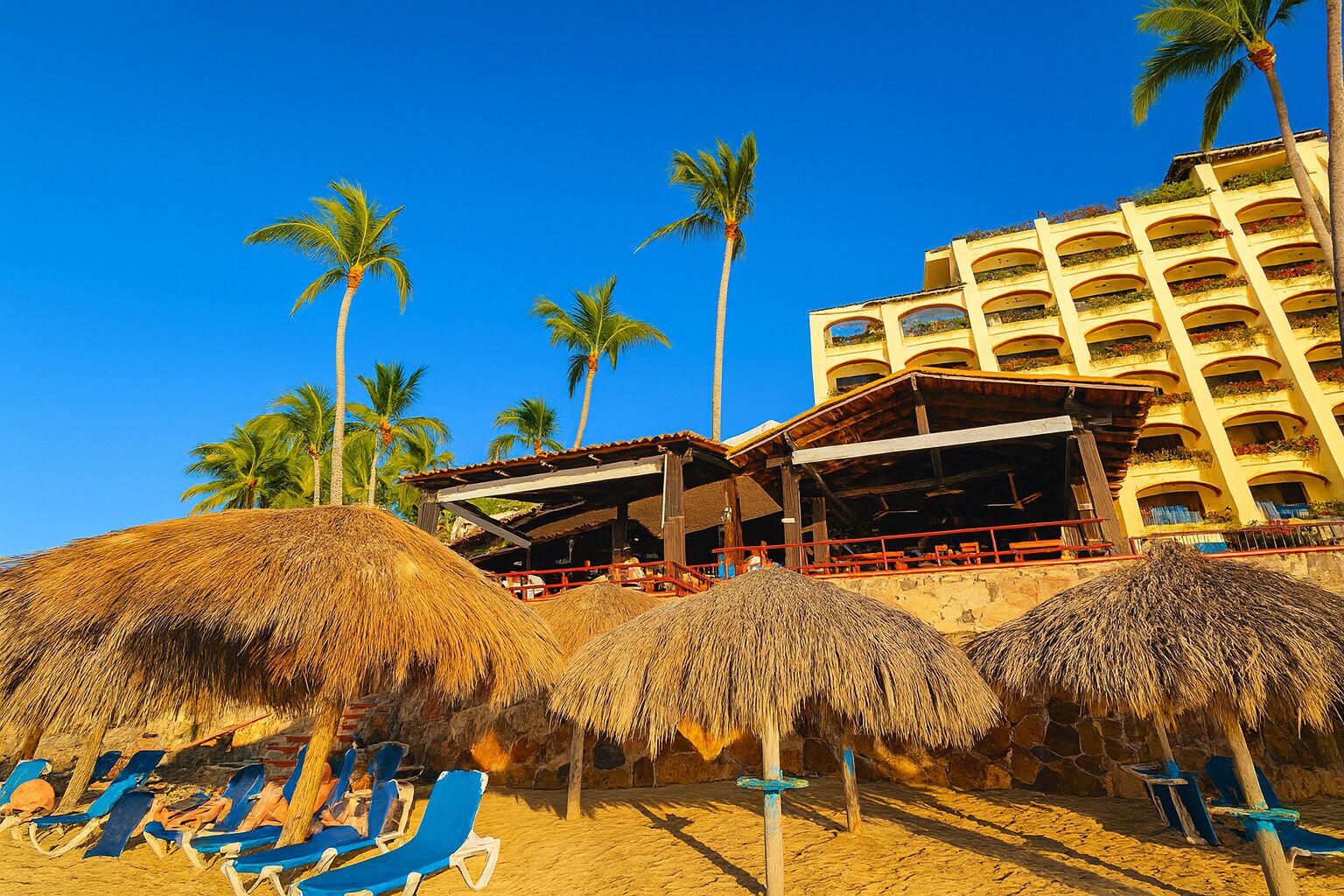 A beachside hotel with thatched umbrellas, blue lounge chairs, and a multi-story building with balconies.