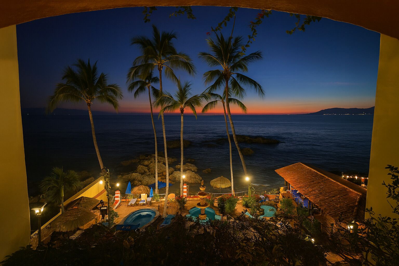 A resort with a pool area, palm trees, and a view of the ocean at dusk.