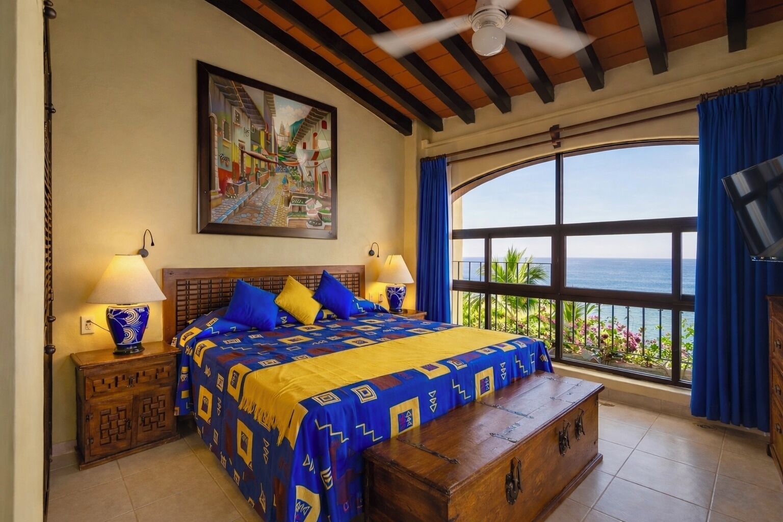 Un dormitorio con una cama grande, dos mesitas de noche con lámparas, un cofre de madera, un ventilador de techo, un ventanal con vista al mar y un cuadro en la pared.