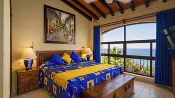 Un dormitorio con una cama grande, dos mesitas de noche con lámparas, un cofre de madera, un ventilador de techo, un ventanal con vista al mar y un cuadro en la pared.