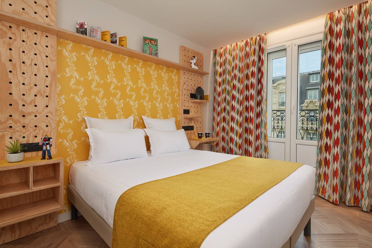 A neatly arranged bedroom with a bed, a shelf with books and decorative items, and a window with curtains.