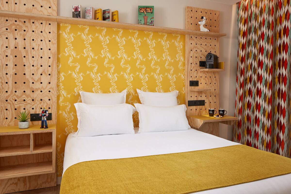 A hotel room with a large bed, wooden headboard, and a yellow patterned wall. There are shelves with books and a small plant.