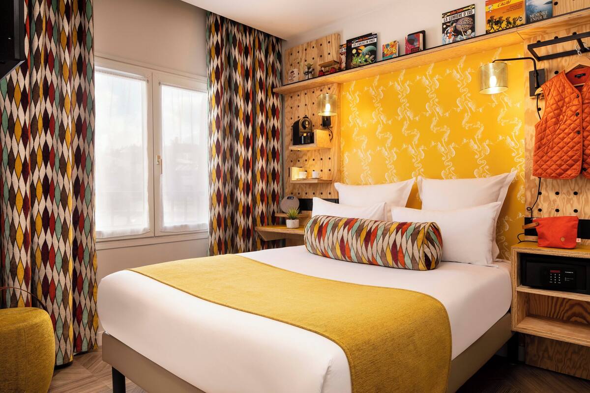 A hotel room with a bed, a bedside table, a lamp, a shelf with books, and a window with curtains.