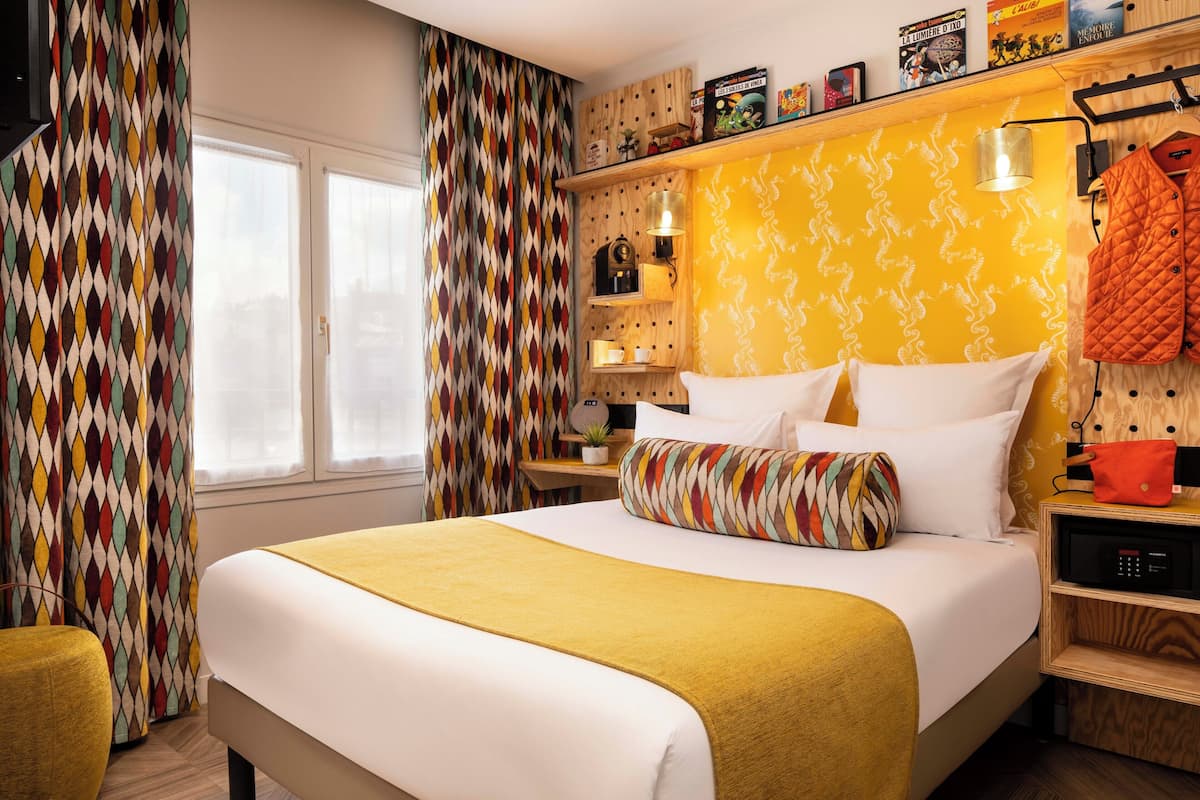 Hotellihuone, jossa on sänky, yöpöytä, lamppu ja hylly, jossa on kirjoja ja takki.