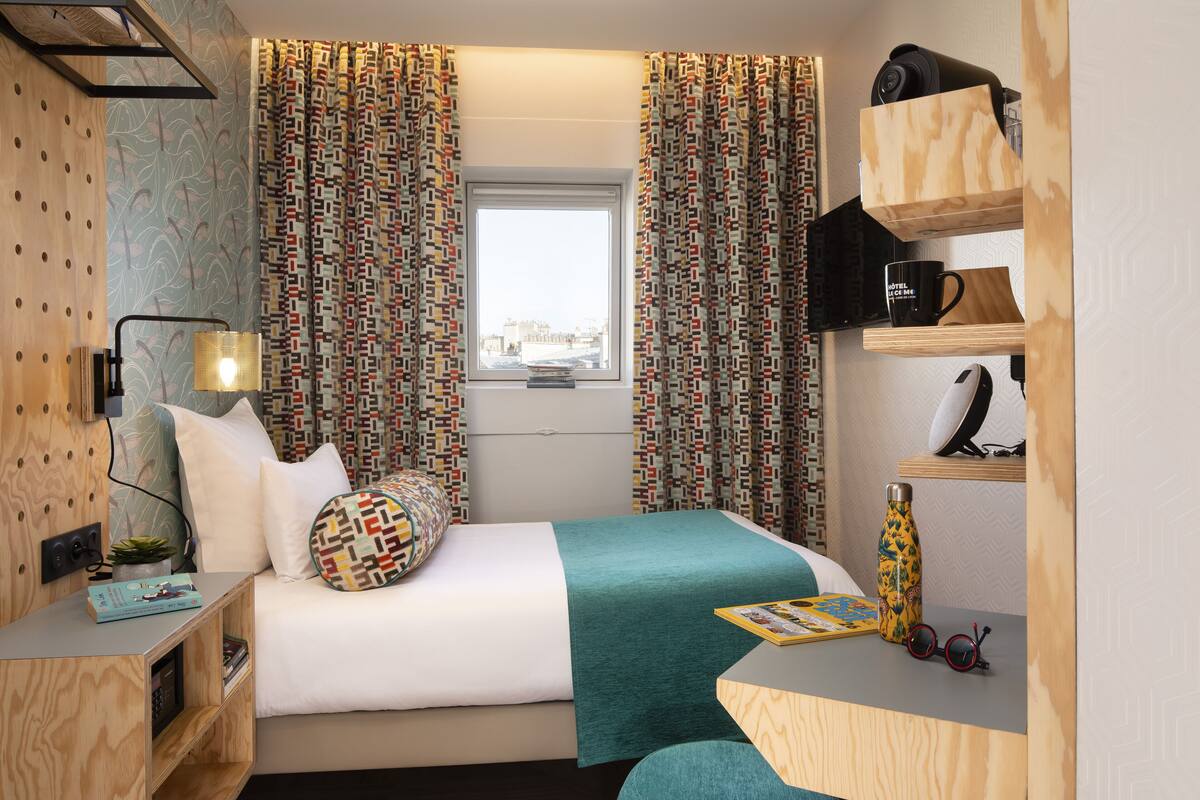A neatly arranged hotel room with a bed, a desk, a shelf with a coffee maker, and a window with patterned curtains.