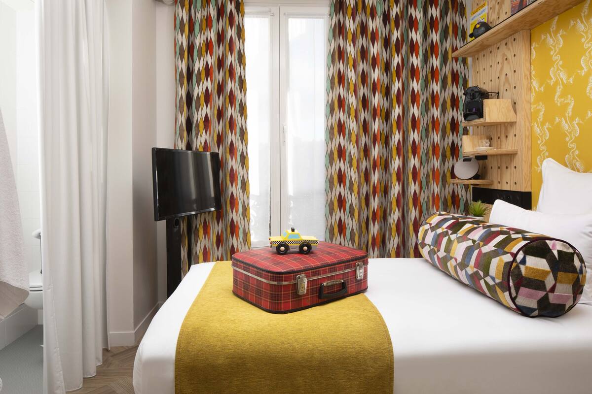 A hotel room with a bed, a suitcase, a toy truck, a television, and a patterned curtain.