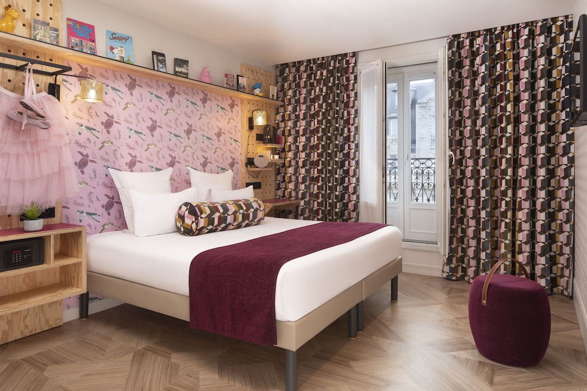A bedroom with a bed, a wooden nightstand, a small table, a purple ottoman, and patterned curtains.