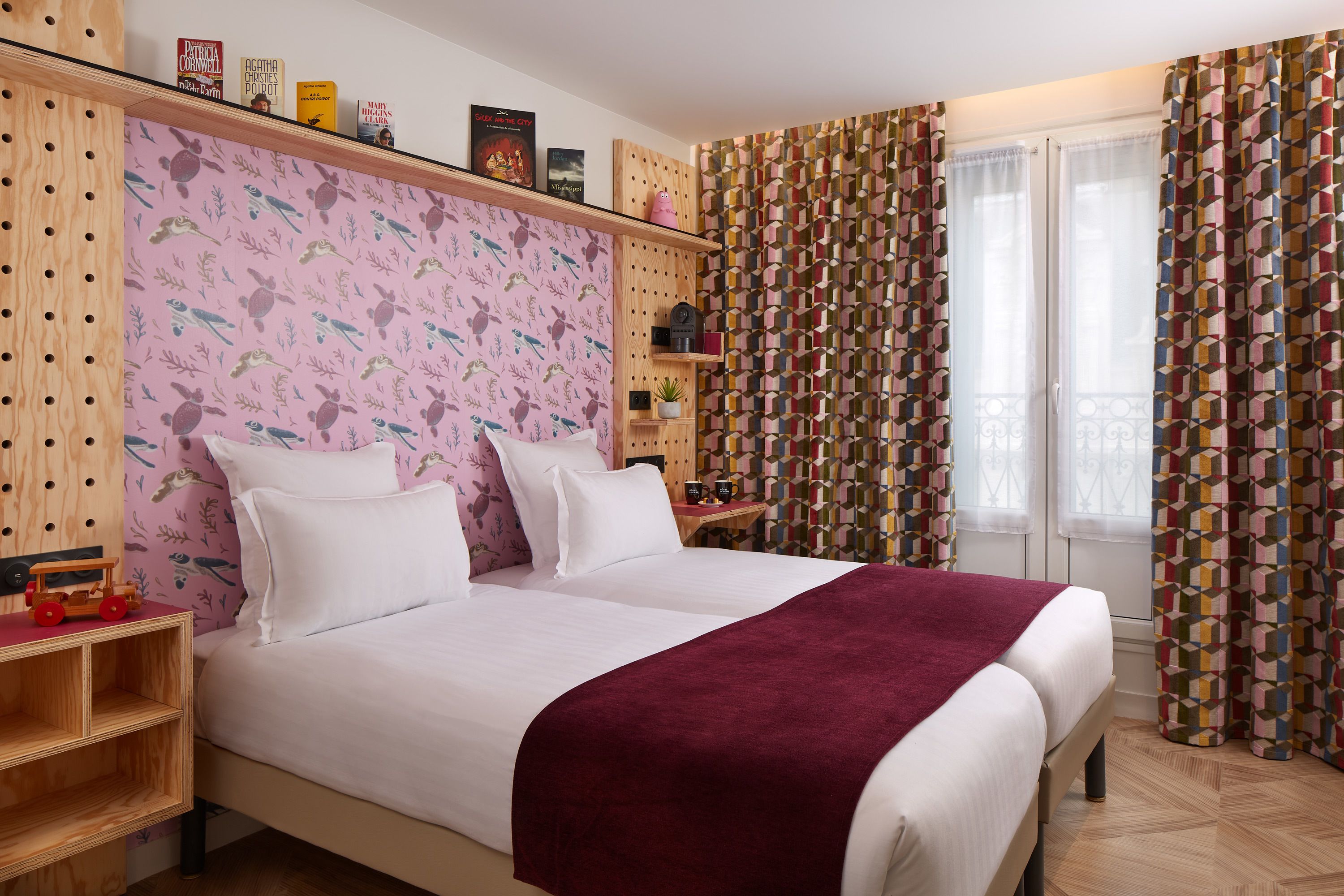 A bedroom with a bed, wooden headboard, a nightstand, a shelf with books, a small table with a plant, and patterned curtains.
