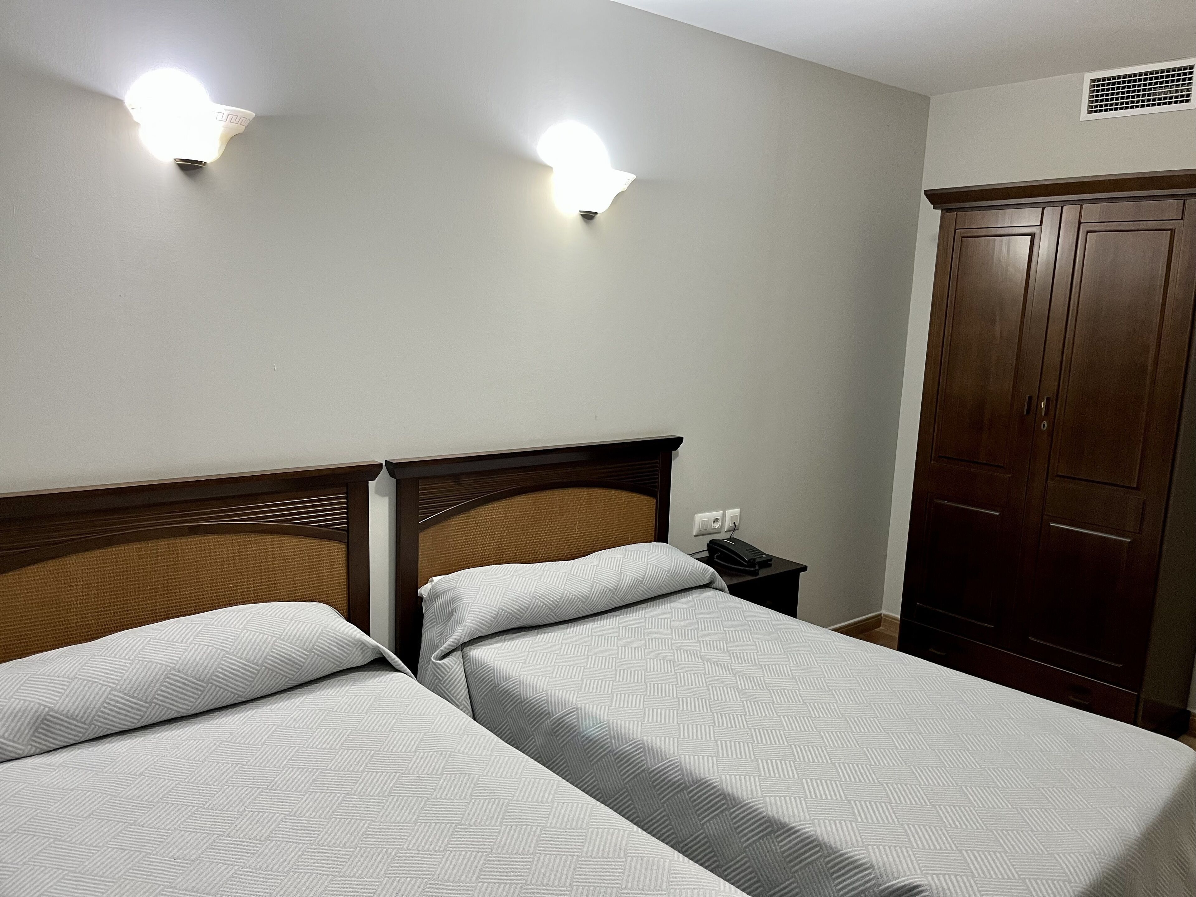 A hotel room with two beds, a wooden headboard, a wardrobe, and two wall-mounted lights.