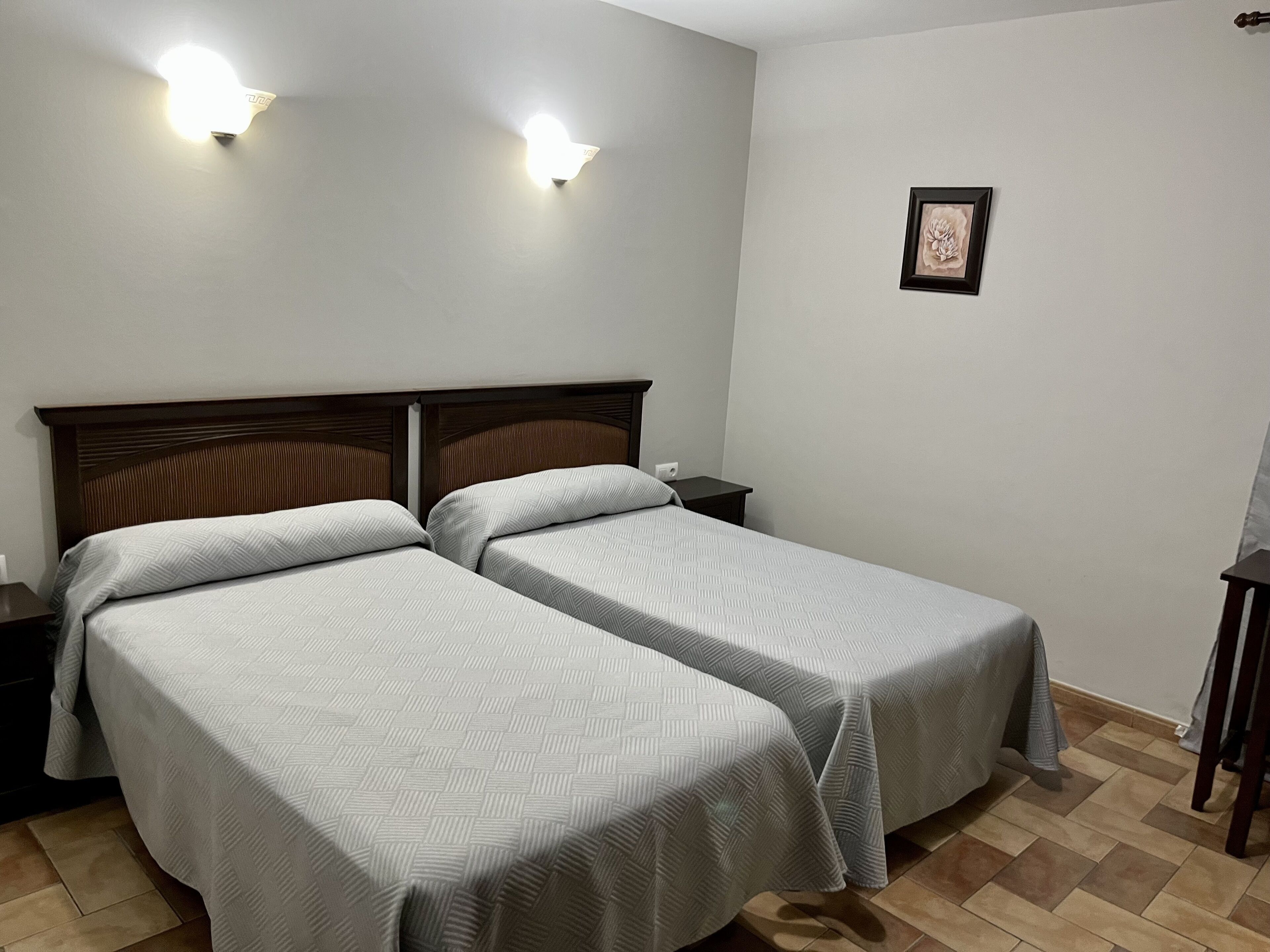 A hotel room with two single beds, a nightstand, a bedside table, a framed picture on the wall, and a ceiling light fixture.