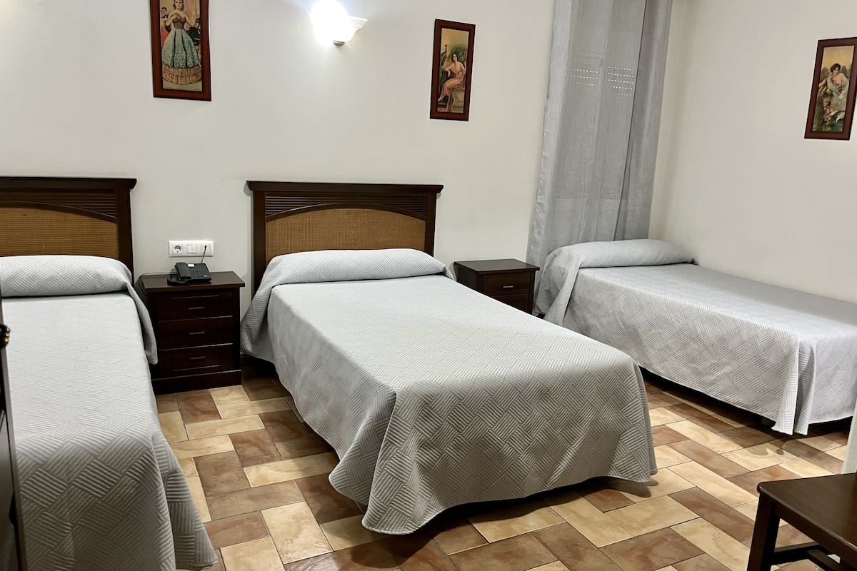 A hotel room with two single beds, a wooden nightstand, a small table, and a wall-mounted light.