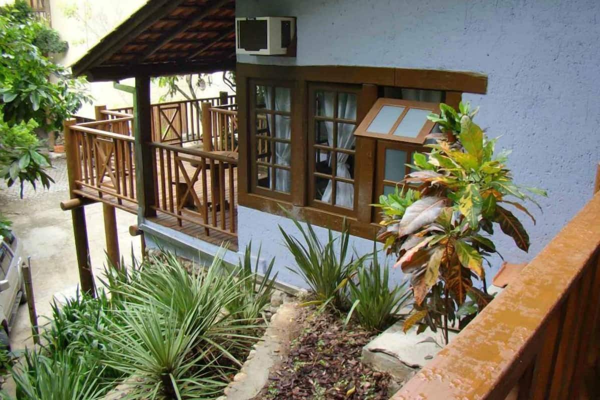 Une terrasse en bois avec une rambarde, un bâtiment blanc doté de fenêtres et diverses plantes.