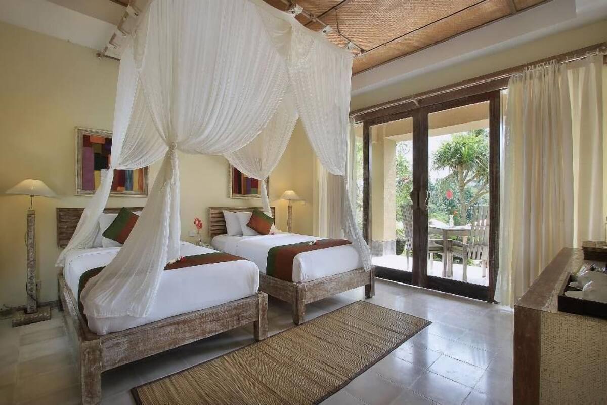 Une chambre avec deux lits en bois, un lit à baldaquin, une grande fenêtre et une vue sur l’extérieur.