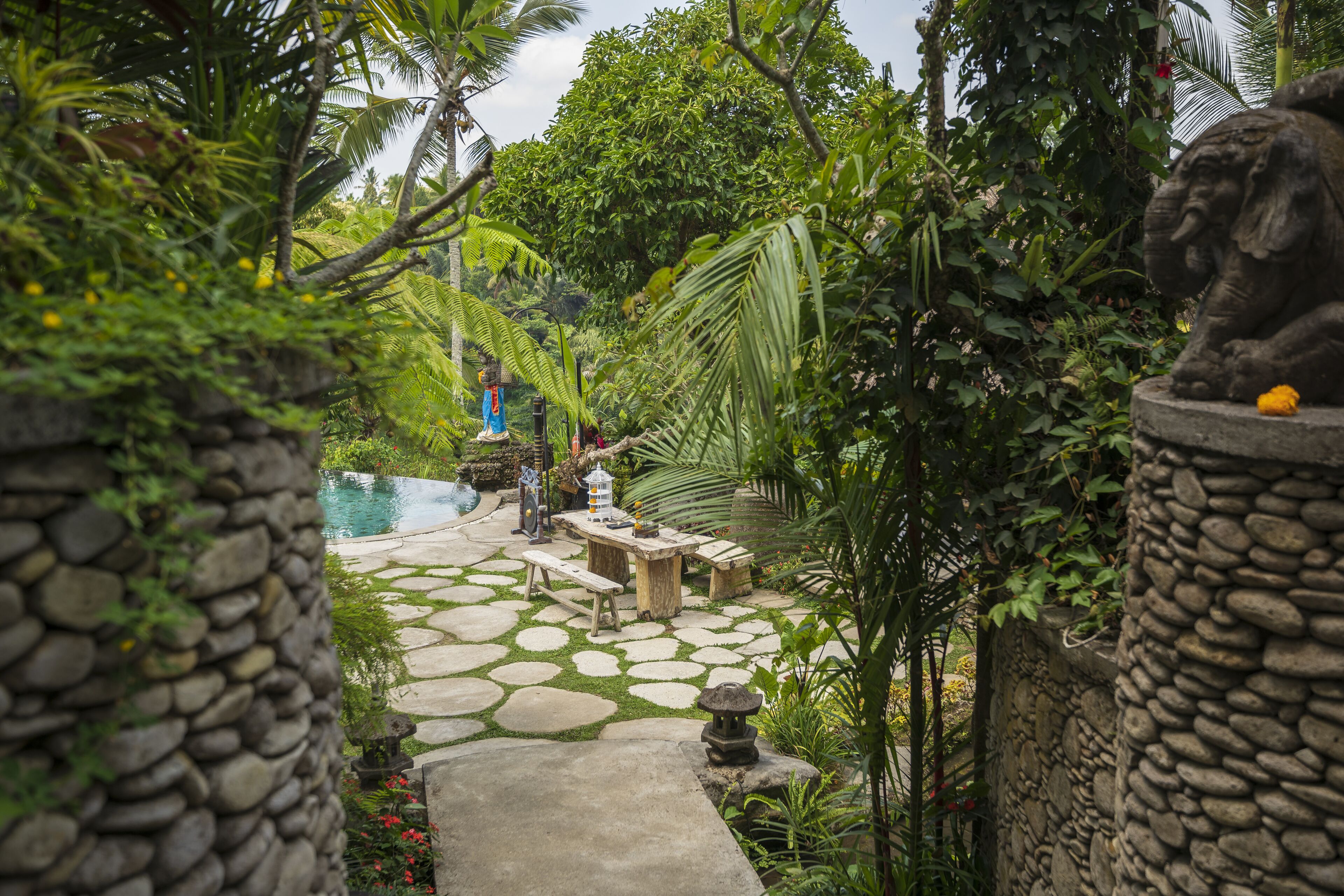 A stone pathway leading to a pool area with a statue of an elephant, surrounded by lush greenery and palm trees.
