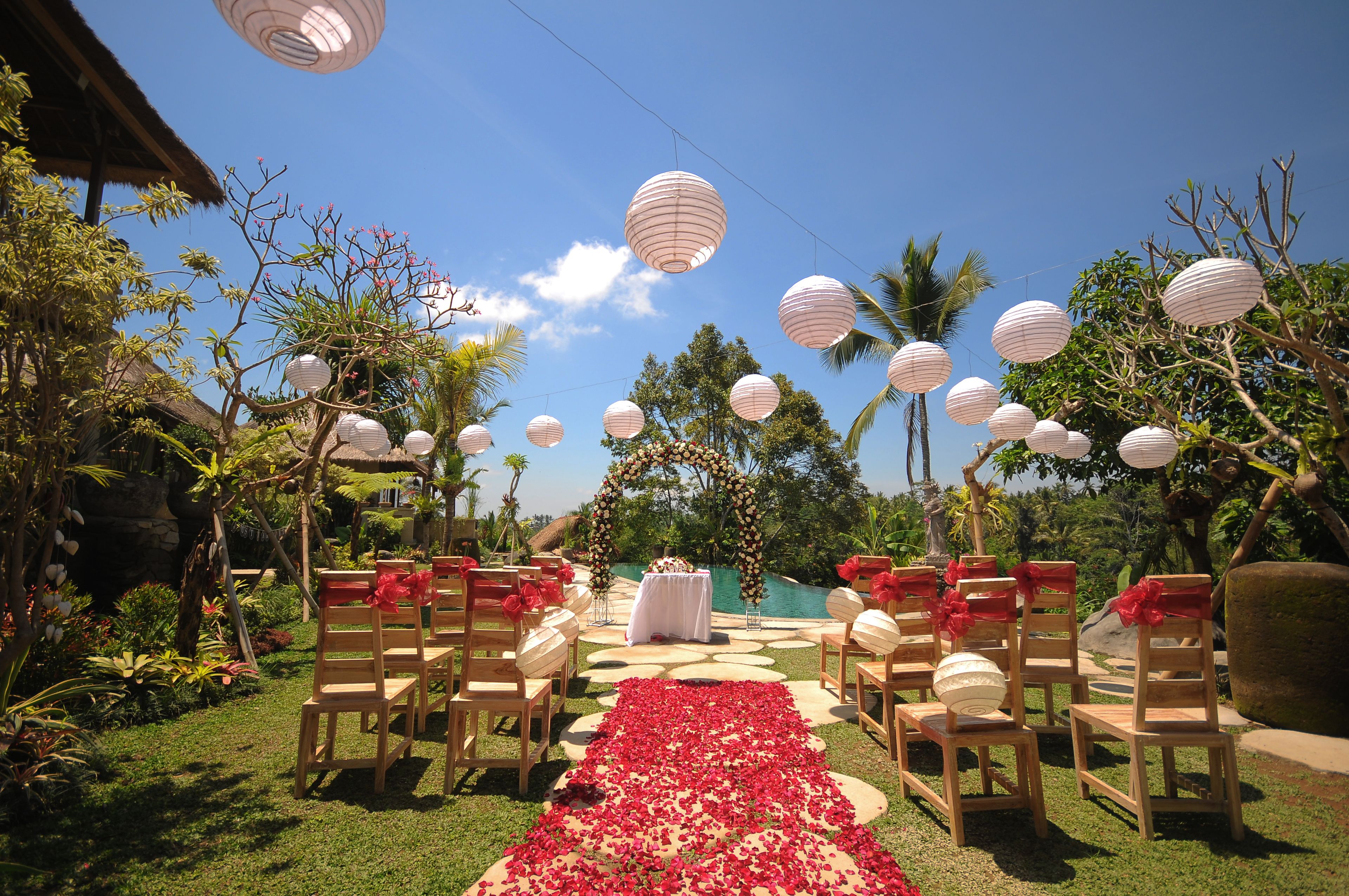 An outdoor wedding setup with wooden chairs, a red carpet, and white paper lanterns.