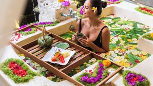Eine Frau in einer mit Blumen und Früchten gefüllten Badewanne, umgeben von einem Holztablett mit Teekanne und Speisen.
